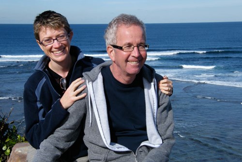 Caroline and Avner sitting together by the ocean, smiling with their arms around each other.