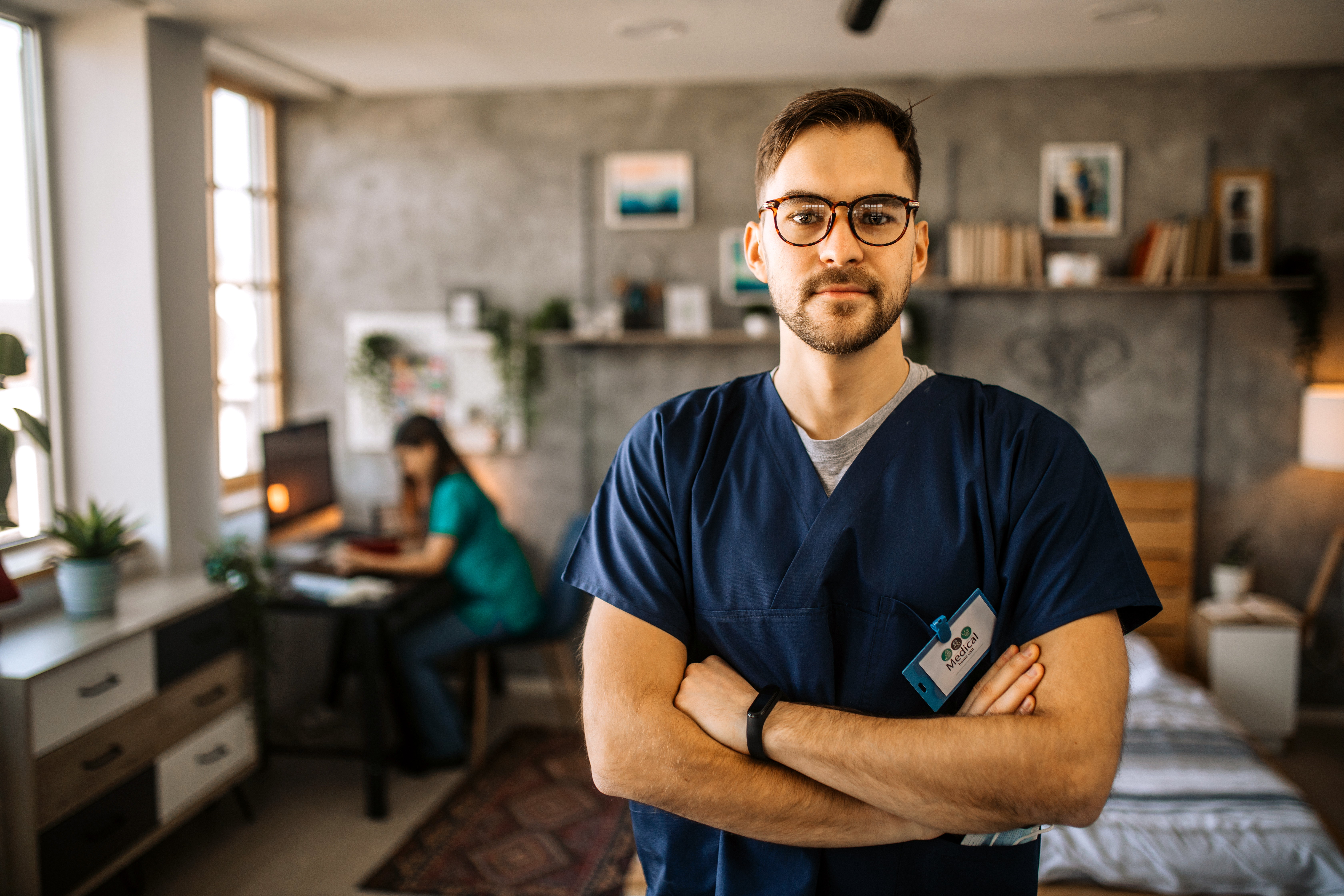 A male healthcare professional in navy blue scrubs stands confidently with arms crossed in a cozy, well-lit room. Behind him, another medical worker is seated at a desk working on a computer. The space has homey decor, including bookshelves, framed art, and natural light streaming through large windows.