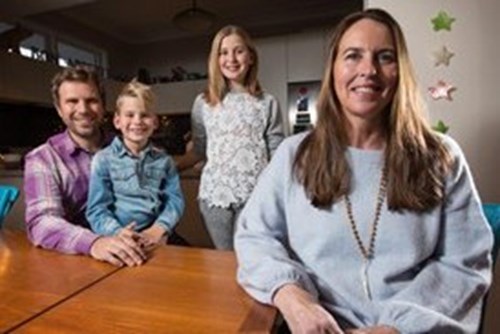 Kristin sits at a table with her husband, son, and daughter smiling behind her in a warmly lit home.