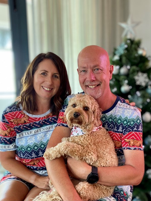 Mark, a man living with pancreatic cancer, sits smiling beside his partner in front of a Christmas tree, both wearing matching festive pyjamas. Mark is holding a fluffy tan-coloured dog. The image reflects warmth, joy, and the support of loved ones during the holiday season.